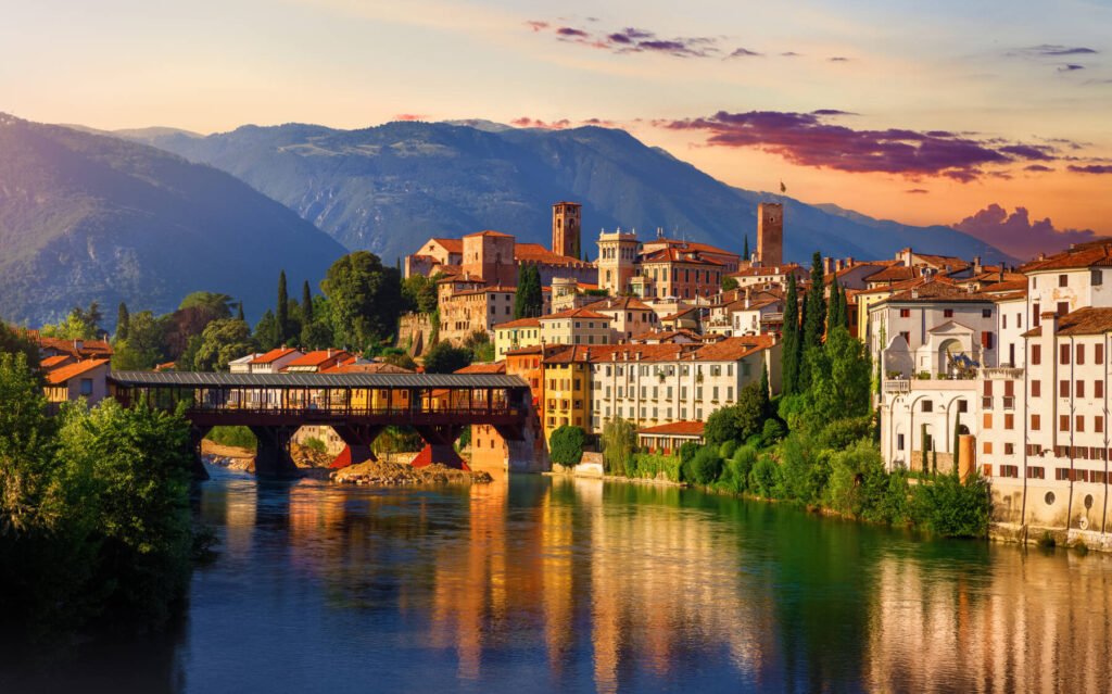 Bassano del Grappa, Veneto, Italy. Bridge Ponte degli Alpini at river Brenta. Panoramic view at old town with vintage building tower, wooden bridge at Alpine mountains scenic sunset landscape. Veneto